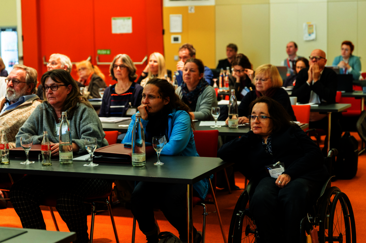Eine Gruppe von Menschen sitzt aufmerksam in einem Konferenzraum mit orangefarbenem Teppichboden und einem hellen, modernen Interieur. Die Teilnehmer blicken nach vorne, offenbar einem Vortrag oder einer Präsentation folgend. Vor ihnen stehen Tische mit Getränkeflaschen, Gläsern und Unterlagen. Unter den Anwesenden befindet sich eine Frau im Rollstuhl im Vordergrund, die konzentriert zuhört. Im Hintergrund sind weitere Teilnehmer zu sehen, die eine Vielfalt an Alter und Stil repräsentieren.