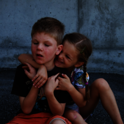 Ein Junge mit kur-zen, hellbraunen Haaren sitzt vor einer Betonwand auf Asphalt. Seine Gesichtszüge zei-gen eine Behinde-rung, und seine Haut weist sichtba-re Rötungen auf. Ein Mädchen mit langen, braunen Haaren, die zu ei-nem seitlichen Zopf geflochten sind, umarmt ihn von hinten und lächelt fröhlich. Sie trägt eine bunte, gemusterte Klei-dung und eine Haarspange. Die Szene wirkt herz-lich und liebevoll.