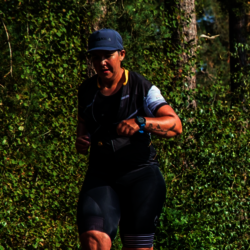 Eine Frau trägt eine blaue Kappe und läuft in sportlicher Kleidung durch einen Wald. Sie hat eine schwarze Laufhose und ein passendes Oberteil mit gelben Akzenten an. An ihrem rechten Arm ist ein Tattoo zu erkennen, und an ihrem linken Handgelenk trägt sie eine Sportuhr. Der Hintergrund zeigt grüne Bäume und Büsche, die von Sonnenlicht durchdrungen sind.