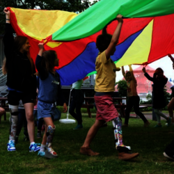 Kinder spielen draußen auf einer Wiese unter einem großen bunten Schwungtuch. Eini-ge der Kinder tra-gen bunte Orthe-sen an den Beinen. Im Hintergrund sind Holzbänke, Gebäude und Bäume zu sehen. Die Szene wirkt lebhaft und fröhlich.