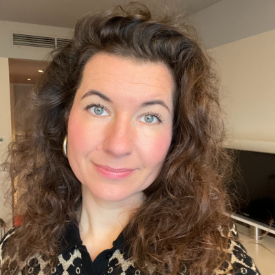 Porträt einer Frau mit schulterlangen, lockigen braunen Haaren. Sie trägt ein gemustertes Oberteil und goldene Creolen, während sie in einem Innenraum steht. Der Hintergrund zeigt unscharf Möbel und eine helle Wand.