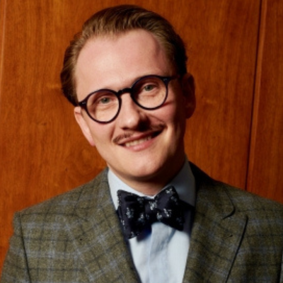 Porträt eines Mannes mit kurzen, zurückgekämmten Haaren, rundlicher Brille und gepflegtem Schnurrbart. Er trägt ein kariertes Sakko, ein hellblaues Hemd und eine schwarze Fliege mit dezentem Muster. Der Hintergrund ist aus Holzpaneelen und verleiht dem Bild eine warme Atmosphäre.