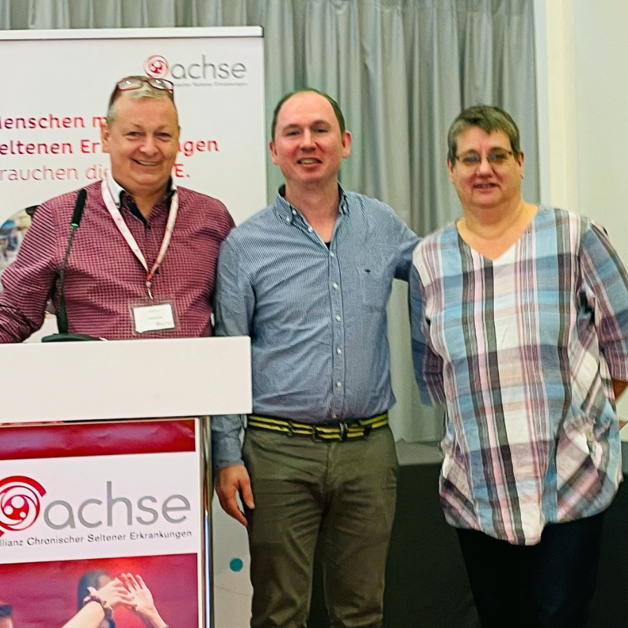 Drei Personen stehen nebeneinander und lächeln in die Kamera. Links steht eine Person mit einer rot-karierten Hemdbluse, einer Namenskarte und einer Brille auf dem Kopf hinter einem Rednerpult mit dem Logo der "ACHSE" (Allianz Chronischer Seltener Erkrankungen). Die mittlere Person trägt ein blau-weiß gestreiftes Hemd und eine beige Hose mit einem gelben Gürtel. Rechts steht eine Person in einer bunt karierten Bluse. Der Hintergrund zeigt einen Vorhang und ein Banner mit ACHSE-Logo.