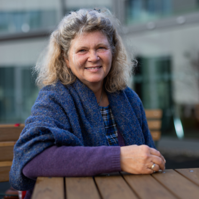 Eine Frau mit lockigem, blonden Haar sitzt an einem Holztisch im Freien. Sie trägt einen blauen Schal und einen lila Pullover. Im Hintergrund ist ein modernes Gebäude mit Glasfenstern unscharf zu erkennen. Die Frau lächelt freundlich in die Kamera.