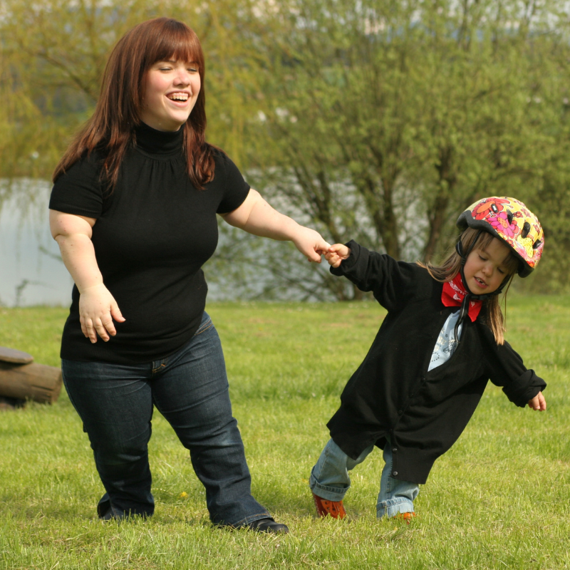 Eine Frau und ein kleines Kind, die beide mit Kleinwuchs leben, halten sich an den Händen und laufen fröhlich auf einer Wiese. Die Frau trägt ein schwarzes Oberteil und Jeans, das Kind einen schwarzen Helm mit buntem Muster, einen roten Schal und eine schwarze Jacke.