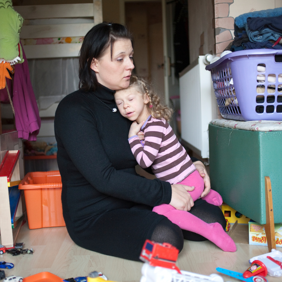 Eine Frau sitzt in einem Kinderzimmer auf dem Boden und hält ein kleines Mädchen mit blonden, zu Locken geformten Haaren in einem gestreiften Pullover und pinken Strumpfhosen liebevoll im Arm. Das Mädchen wirkt erschöpft und ruht sich an der Schulter der Frau aus. Im Hintergrund sind ein Etagenbett, ein Wäschekorb und Spielsachen zu sehen.