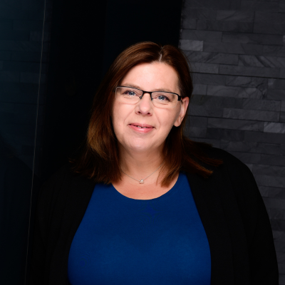 Porträtfoto einer Frau mit schulterlangem, dunkelbraunem Haar. Sie trägt eine Brille mit schwarzem Rahmen, ein blaues Oberteil und eine schwarze Jacke. Der Hintergrund besteht aus einer modernen, dunklen Steinwand.