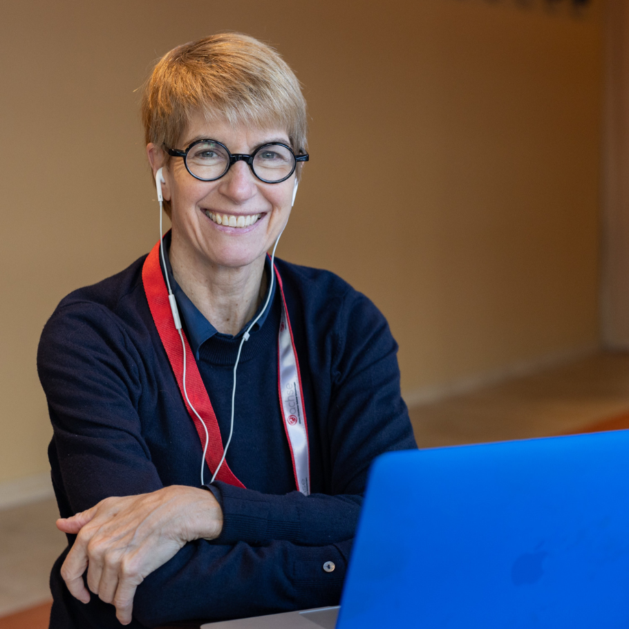 Eine Frau mit kurzen blonden Haaren und runder Brille sitzt lächelnd vor einem blauen Laptop. Sie trägt ein dunkelblaues Oberteil und ein rotes Schlüsselband. Weiße Kopfhörer sind in ihre Ohren gesteckt. Im Hintergrund ist eine schlichte, helle Wand sichtbar.
