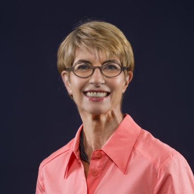 Eine Frau mit kurzen blonden Haaren und runder Brille lächelt in die Kamera. Sie trägt eine korallfarbene Bluse mit Kragen. Der Hintergrund ist dunkelblau und einfarbig gehalten.