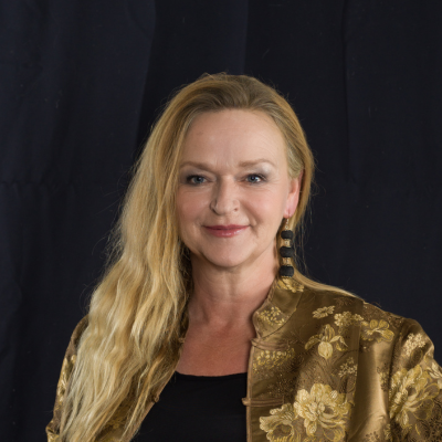 Portrait einer Frau mit langen, welligen, blonden Haaren. Sie trägt eine goldene Jacke mit floralem Muster und lange schwarze Ohrringe. Ihr Lächeln ist freundlich und zugewandt. Der Hintergrund ist dunkel.