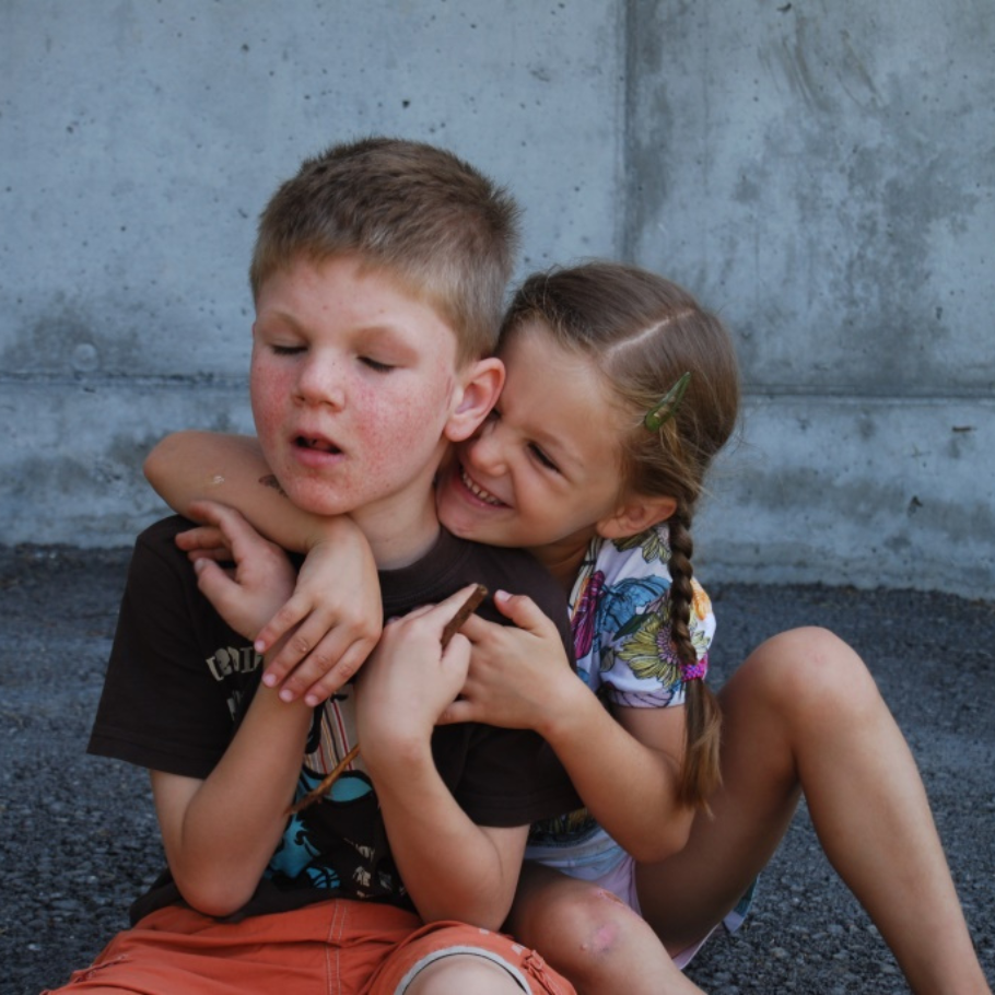 Ein Junge mit kur-zen, hellbraunen Haaren sitzt vor einer Betonwand auf Asphalt. Seine Gesichtszüge zei-gen eine Behinde-rung, und seine Haut weist sichtba-re Rötungen auf. Ein Mädchen mit langen, braunen Haaren, die zu ei-nem seitlichen Zopf geflochten sind, umarmt ihn von hinten und lächelt fröhlich. Sie trägt eine bunte, gemusterte Klei-dung und eine Haarspange. Die Szene wirkt herz-lich und liebevoll.