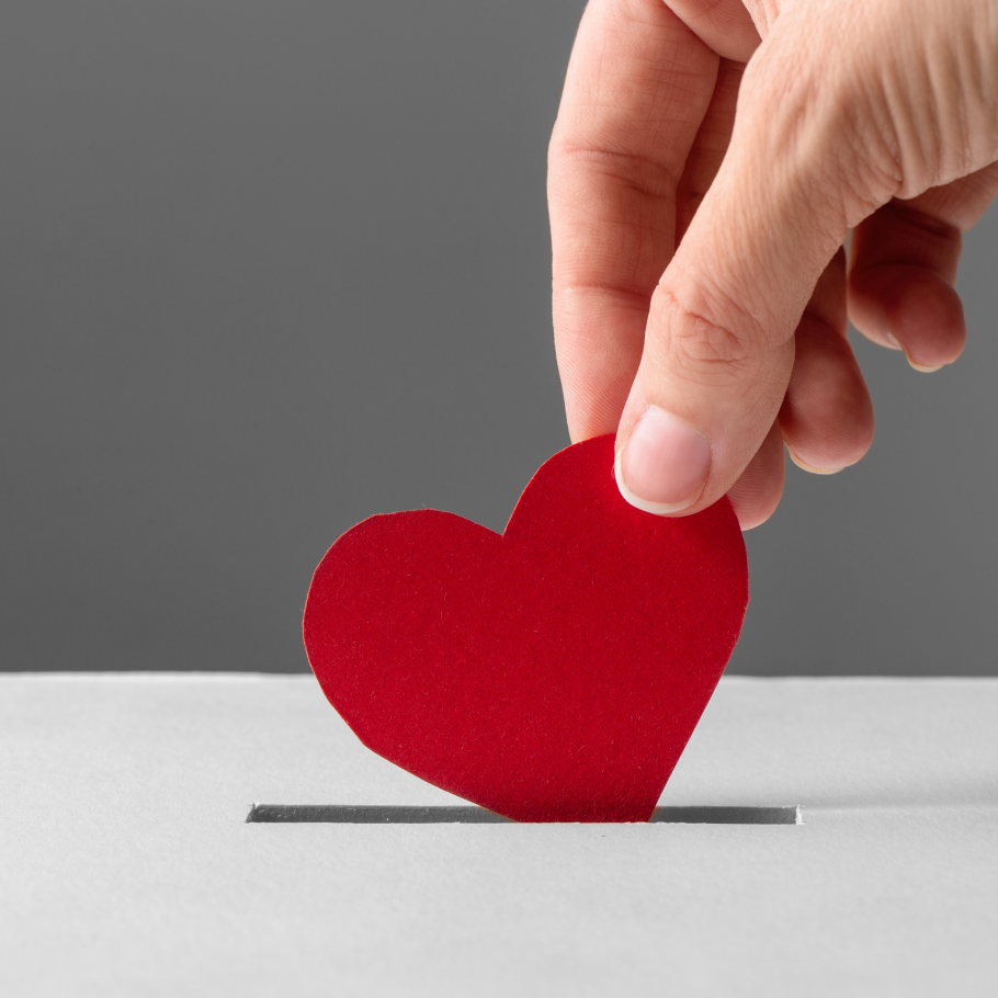 Eine Hand hält ein rotes, herzförmiges Stück Papier und steckt es in einen schmalen Schlitz, der wie ein Spendenkasten wirkt. Der Hintergrund ist grau, und die Szene symbolisiert eine Spende aus Herzen.