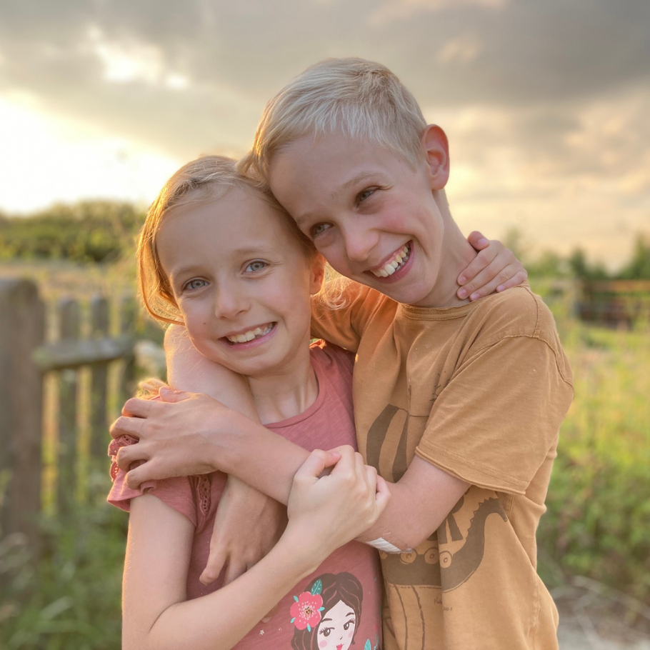 Ein Mädchen mit weggesteckten, langen blonden Haaren und ein Junge mit kurzen blonden Haaren umarmen sich draußen in einer ländlichen Umgebung bei Sonnenuntergang und lächeln in die Kamera. Bei genauerem Hinsehen ist erkennbar, dass der Junge eine Behinderung hat. Im Hintergrund sind ein Holzzaun und eine Wiese zu sehen, die in warmem Licht erstrahlen.